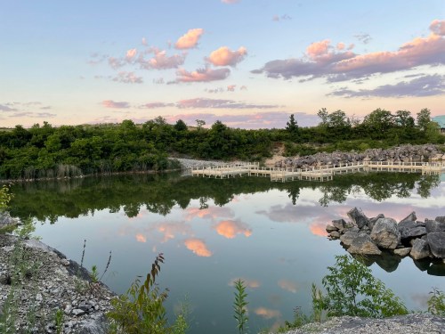 A serene landscape featuring a calm pond reflecting clouds and trees, surrounded by rocky terrain and greenery.