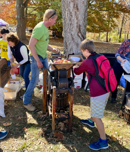 A man and a child use an apple press outdoors, surrounded by autumn trees and people enjoying a fall festival.