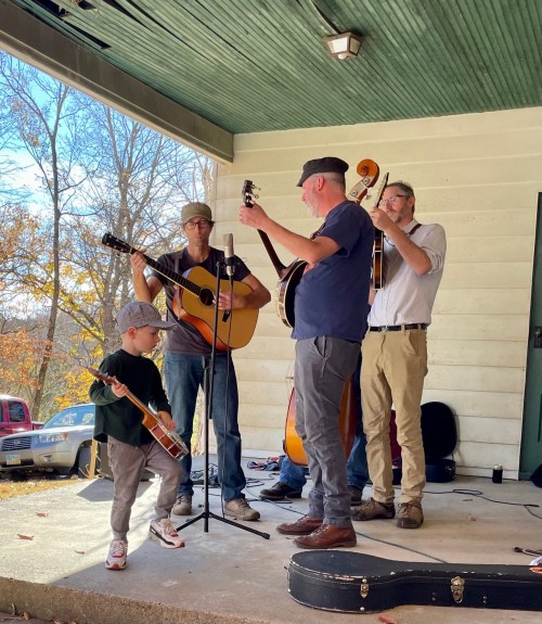 A group of musicians performs on a porch, with a child playing a small guitar alongside them.