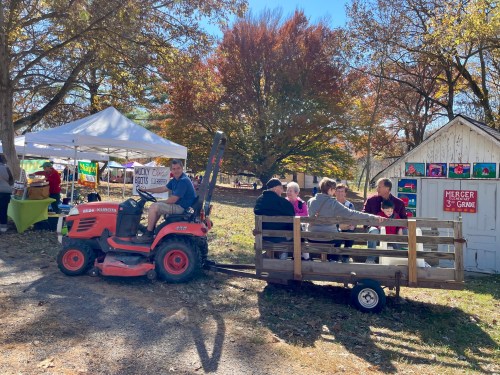 A tractor pulls a wooden wagon with people seated, near tents and a small shed in a colorful autumn setting.