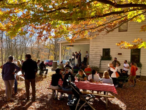 A gathering of people outdoors in autumn, enjoying food and conversation under colorful trees near a white building.