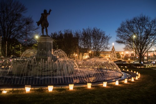 A fountain with illuminated candles at dusk, featuring a statue and a Christmas tree in the background.