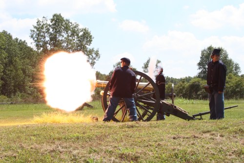 A historical reenactment of soldiers firing a cannon in a grassy field under a blue sky.