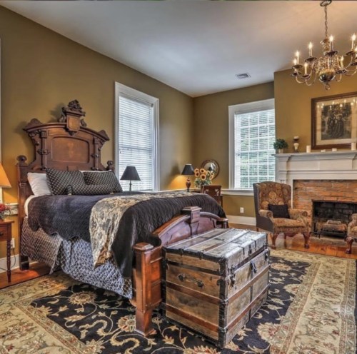 Cozy bedroom with a dark wood bed, vintage trunk, and a fireplace, featuring warm tones and elegant decor.