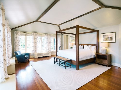 Spacious bedroom with a four-poster bed, large windows, and a cozy armchair, featuring wooden accents and soft decor.