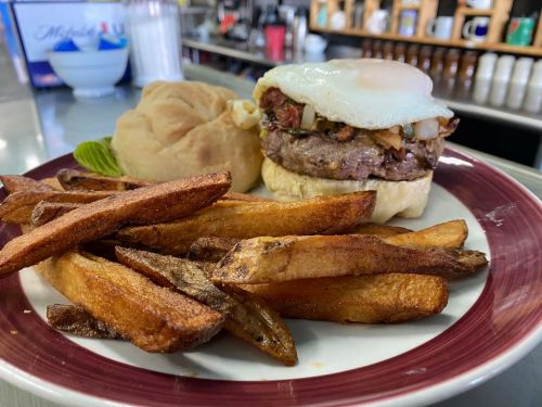 A plate with a burger topped with an egg, served with fries and a soft bun, in a diner setting.