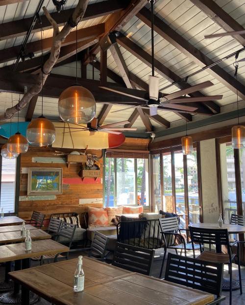Cozy restaurant interior with wooden beams, pendant lights, and surfboards, featuring tables and a relaxed atmosphere.