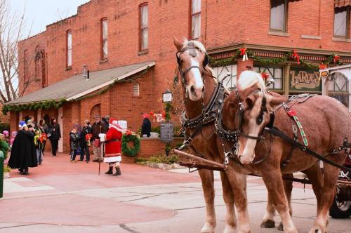 Two horses in harness stand in front of a festive street scene with people in holiday attire and decorations.