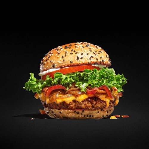 A delicious burger with a sesame seed bun, lettuce, tomato, bacon, and melted cheese against a black background.