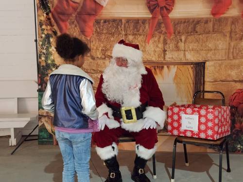 A child in a colorful jacket talks to Santa Claus, who is seated in front of a festive backdrop and a box labeled "Letters to Santa."