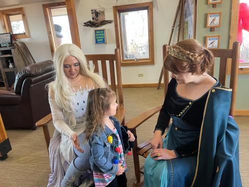 Two women in costumes interact with a young girl in a cozy indoor setting with wooden decor.