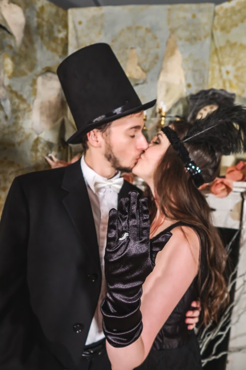 A couple in vintage attire shares a kiss, with the woman wearing a feathered headband and black gloves.