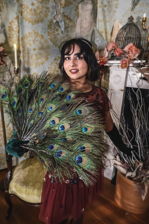 A woman in a vintage dress holds a peacock feather fan, posing elegantly in a richly decorated room.