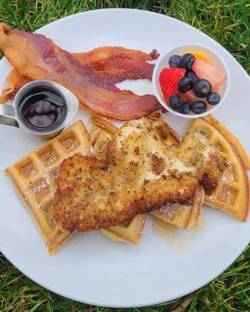 A plate of waffles topped with fried chicken, bacon, and a side of mixed fruit with syrup.