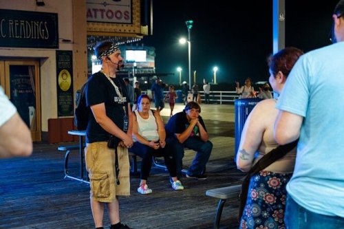 A man speaks to a small group on a boardwalk at night, with people seated nearby. Bright lights illuminate the scene.