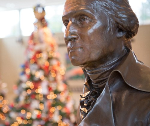 A close-up of a bronze statue of George Washington, with a decorated Christmas tree blurred in the background.