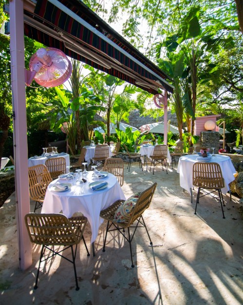Outdoor dining area with white tables, wicker chairs, and lush greenery, featuring pink fans and a sunny atmosphere.