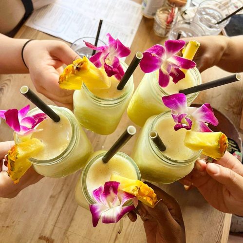 Five tropical drinks with pineapple and orchid garnishes, held by hands around a wooden table.
