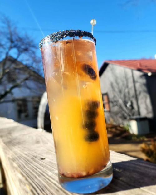 A tall cocktail glass with a dark rim, filled with a colorful drink and garnished with a skewer, set against a clear blue sky.