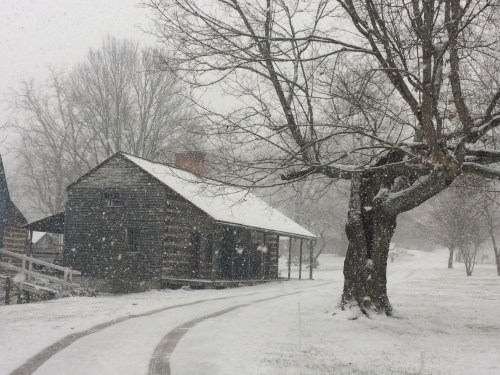 A snowy scene featuring a rustic wooden house surrounded by trees, with snowflakes falling gently.