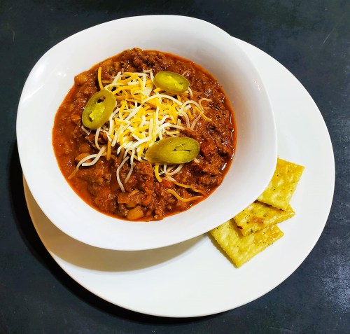 A bowl of chili topped with cheese and jalapeños, served with crackers on a white plate.