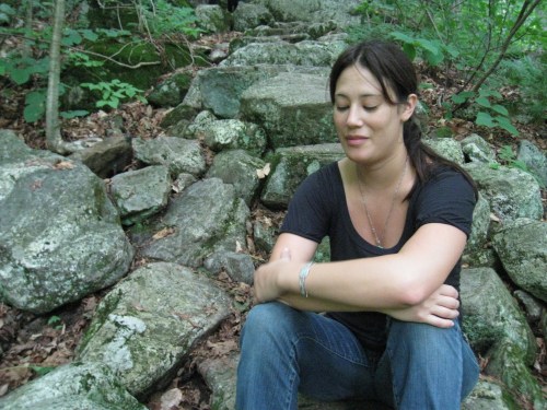 A woman sits on rocky terrain in a forest, eyes closed, with a serene expression amidst greenery and stones.