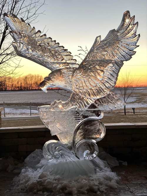 A stunning ice sculpture of an eagle in flight, set against a sunset backdrop with a snowy landscape.
