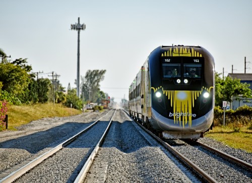 A modern train approaches on a gravel track, surrounded by greenery and utility poles under a clear sky.