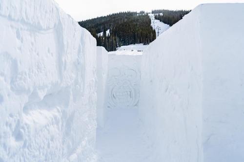 A snowy pathway between tall snow walls, leading to a forested mountain in the background.