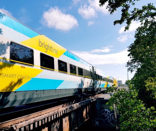 A Brightline train with a blue and yellow design travels over a bridge, surrounded by trees and a clear sky.