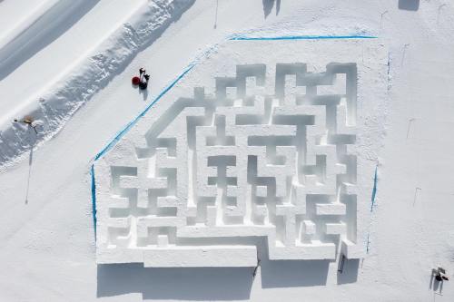 Aerial view of a snow maze with intricate pathways, surrounded by untouched snow and a few people nearby.
