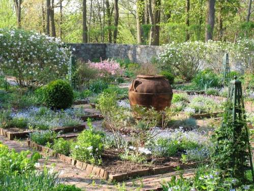 A serene garden with blooming flowers, greenery, and a large clay pot surrounded by stone walls and trees.