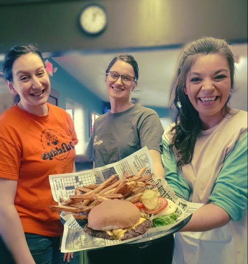 Three smiling women hold a large plate of a burger and fries, showcasing a cheerful dining atmosphere.