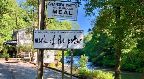 Sign for "Grandpa Watts Water Ground Meal" and "Mark of the Potter" shop near a river, surrounded by greenery.