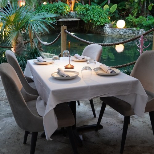 A beautifully set dining table with white tablecloth, surrounded by lush greenery and soft lighting near a pond.