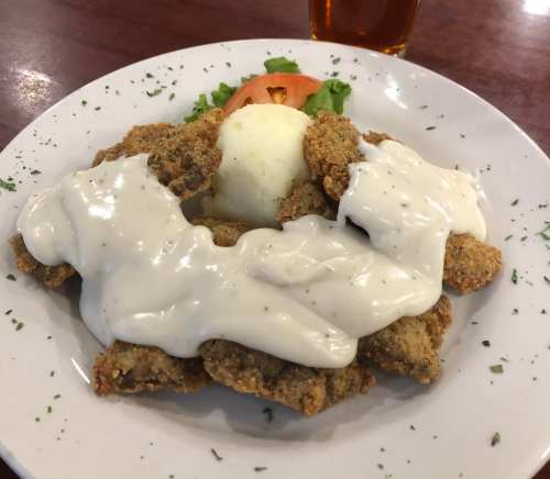 A plate of fried food topped with creamy sauce, accompanied by a salad of lettuce and tomato, with a drink in the background.