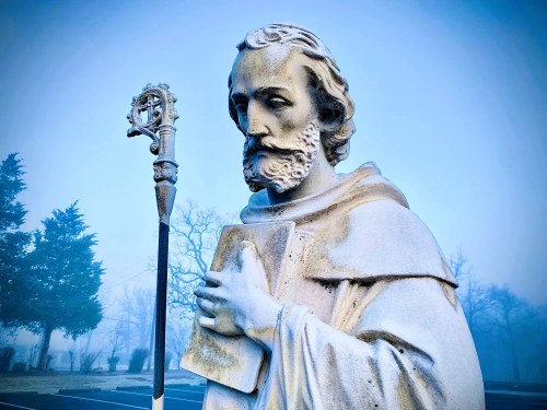 A close-up of a stone statue of a bearded man holding a book, with a staff, set against a foggy background.