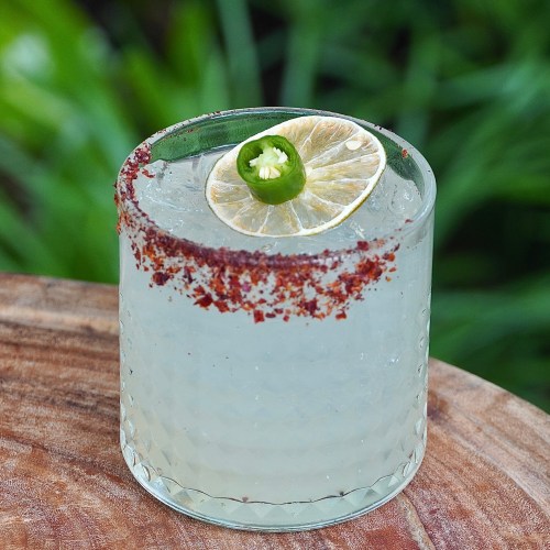 A refreshing cocktail with a salted rim, garnished with a lime slice and a slice of jalapeño, on a wooden surface.