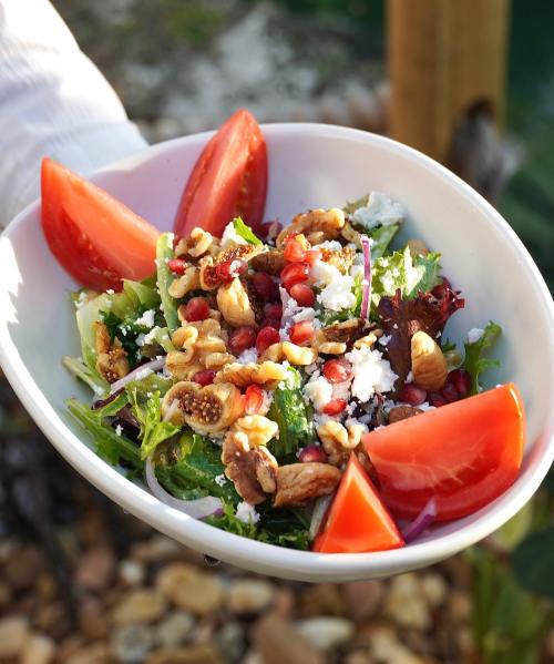 A fresh salad in a white bowl, topped with tomatoes, walnuts, pomegranate seeds, and feta cheese.