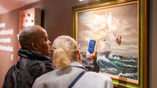 Two people admire a painting of a ship in turbulent waters, one taking a photo with a smartphone.