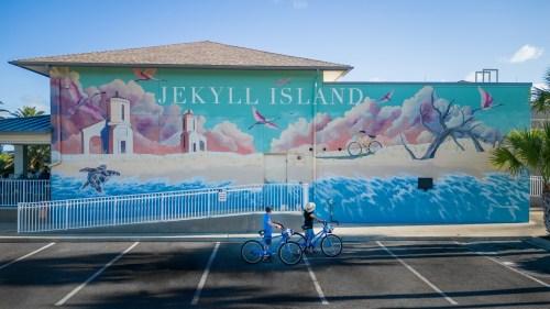 A colorful mural of Jekyll Island featuring birds, water, and cyclists in front of a building.