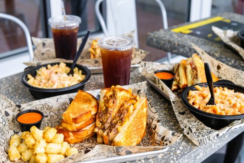 A table with various comfort foods: grilled cheese sandwiches, mac and cheese, tater tots, and iced drinks.