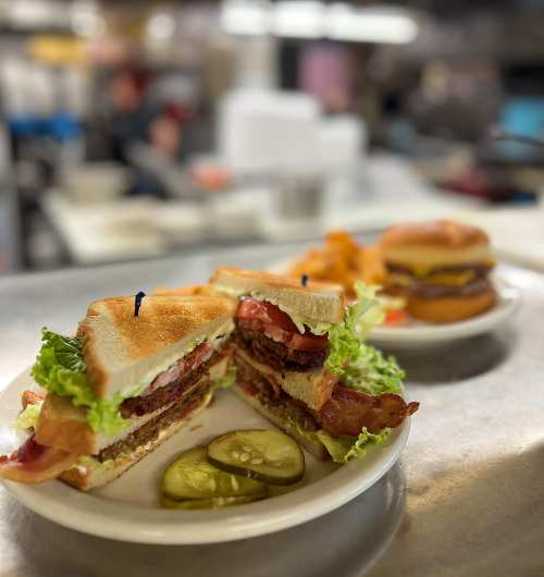 A close-up of a layered sandwich with bacon, lettuce, tomato, and pickles, served on a plate in a diner setting.