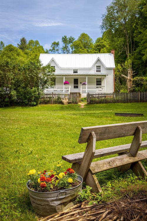 A white farmhouse with a metal roof, surrounded by greenery and flowers, with a wooden bench in the foreground.