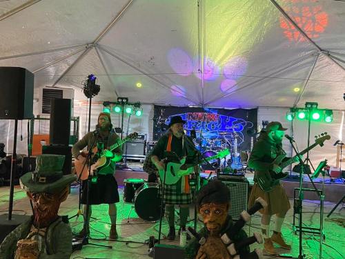 A band performs under a tent with colorful lights, featuring musicians in kilts and decorative statues in the foreground.