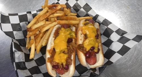 Two loaded hot dogs topped with chili and cheese, served with a side of crispy French fries in a checkered basket.