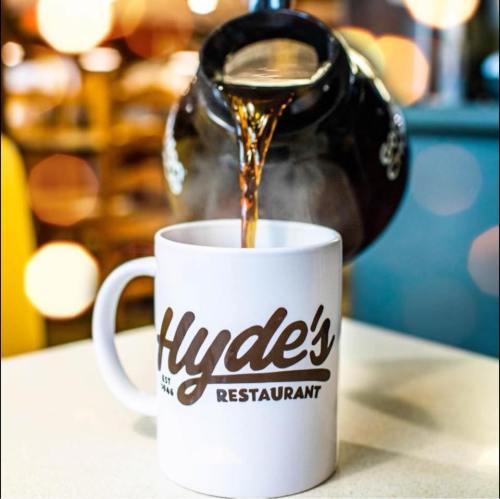 A coffee pot pours coffee into a white mug with "Hyde's Restaurant" printed on it, set against a blurred background.