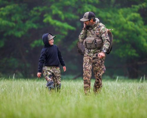 A man and a boy in camouflage outfits walk together through a grassy field, surrounded by green trees.