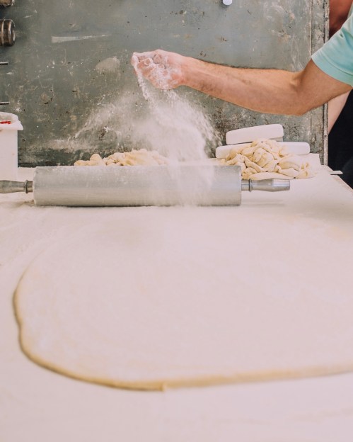 A person sprinkles flour over rolled-out dough on a countertop, with a rolling pin nearby.