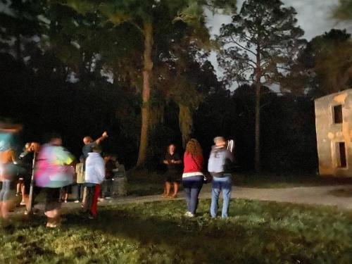 A group of people gathered in a dimly lit forest, some pointing towards a building, surrounded by trees at night.